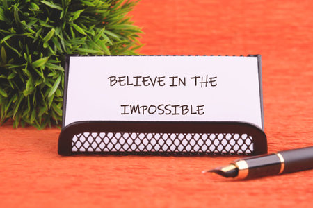 Motivational quote. BELIEVE IN THE IMPOSSIBLE text written on the business card holder. Next to it is a fountain pen and a small ornamental plant on an orange background.の写真素材