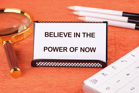 Finance and business concept. Text BELIEVE IN THE POWER OF NOW on the card next to a magnifying glass, pencils and a calculator.の写真素材