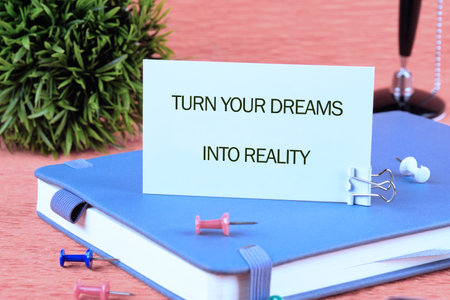 TURN YOUR DREAMS INTO REALITY words written on a card on a blue notebook next to colorful pins and...