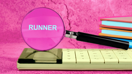 The word RUNNER through a magnifying glass placed on a calculator, and next to it - a stack of colorful books on a bright pink backgroundの写真素材
