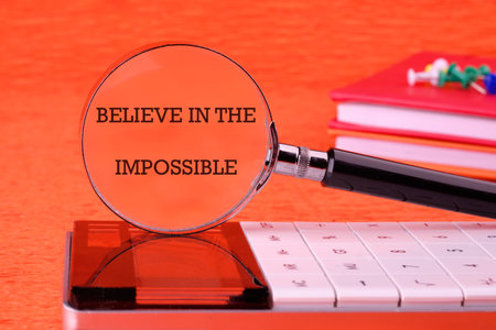 Motivational quote. BELIEVE IN THE IMPOSSIBLE text focused through a magnifying glass on an orange background, based on a calculator.の写真素材