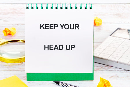 Concept words KEEP YOUR HEAD UP written on a desktop notebook with decorative elements such as a calculator, a magnifying glass and yellow origami.の写真素材