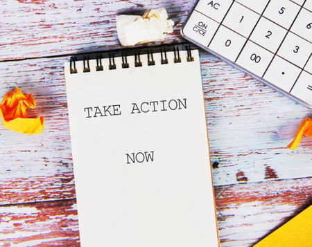 Encouraging motivation, productivity and determination concept. TAKE ACTION NOW written on a blank notebook that lies on a wooden surface next to a calculator and crumpled paper.