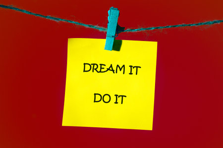 Motivational words DREAM IT, DO IT written on a bright yellow note, which is attached to the shoelace with a blue clothespin on a bright red background.の写真素材