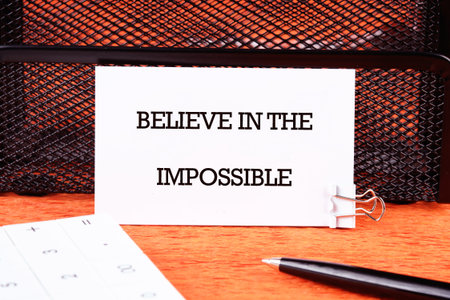 Motivational quote. BELIEVE IN THE IMPOSSIBLE text written on a business card next to a pen and a calculator in a prominent place.の写真素材