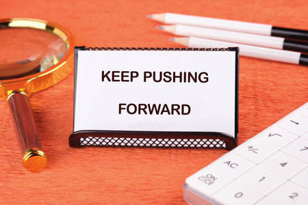 Concept text KEEP PUSHING FORWARD on the card next to a magnifying glass, pencils and a calculator.