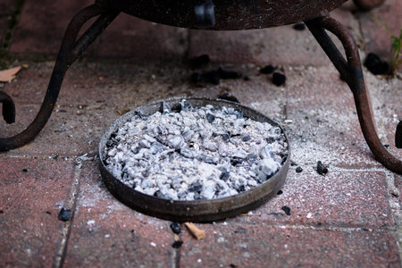 Ashes and charcoal remnants fill a circular tray beneath a fire pit on a brick patio where friends...