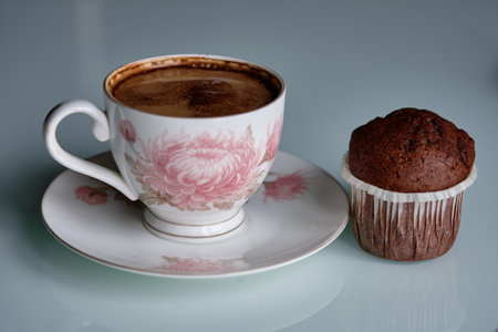 A delicate floral cup holds rich coffee beside a freshly baked chocolate muffin on a white table in...