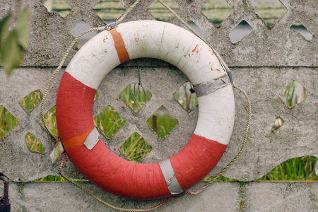 A red and white lifebuoy hangs on a textured wall, with green grass visible in the background,...