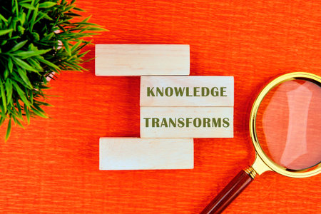 KNOWLEDGE TRANSFORMS concept words assembled from wooden blocks in composition with magnifying glass and green plant on orange background, top view.の写真素材