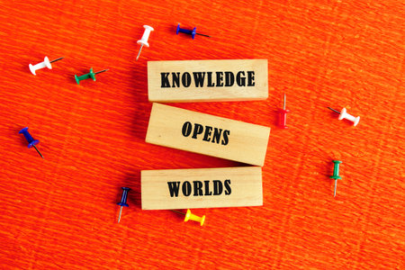 Academic education learning concept. Words KNOWLEDGE OPENS WORLDS assembled from wooden blocks in a composition with stationery pins on an orange provocative background, top view.の写真素材