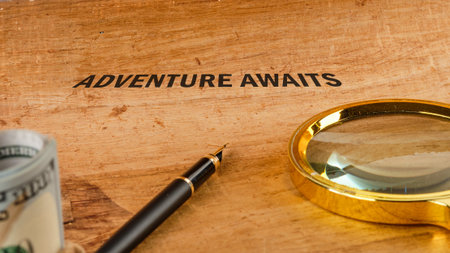 A rolled stack of cash, a sleek pen, and a magnifying glass sit near the words Adventure Awaits etched on a wooden surface, backing travel.の写真素材