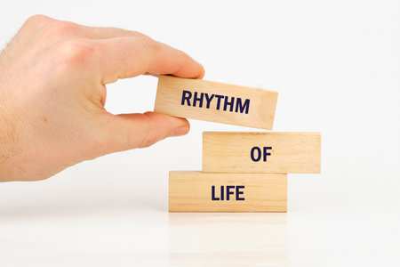 A hand is carefully stacking wooden blocks, each bearing a piece of the phrase RHYTHM OF LIFE. The scene conveys a sense of deliberate construction and balance in the pursuit of well-being.の写真素材