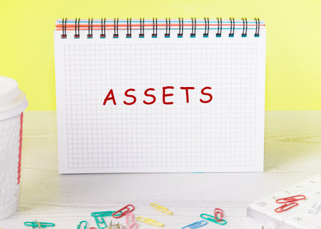 A spiral notebook sits on a desk with the word assets written on the paper. Nearby are scattered paperclips and a coffee cup. The backdrop is light yellow.の写真素材