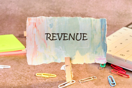 A colorful note with the word REVENUE is displayed with a wooden clothespin. Nearby are paperclips, sticky notes, and a calculator on a textured surface.の写真素材
