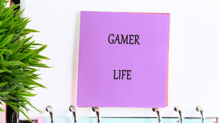 A purple note with GAMER LIFE written in black letters hangs from a ringed notebook. A green plant sits to the left, creating a bright and clean workspace.の写真素材