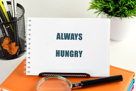 A notebook with the phrase Always Hungry stands on orange books. A magnifying glass rests in front,...