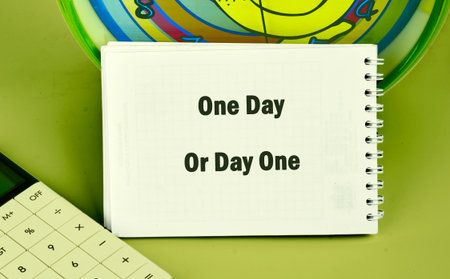 A notepad displaying One Day Or Day One sits near a calculator and a colorful clock, all against a green background, suggesting thoughts about starting goals now.の写真素材