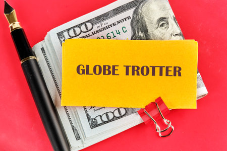 A stack of cash sits with a yellow card that says GLOBE TROTTER, next to a black pen and a red paper clip, set against a bright red backdrop.の写真素材