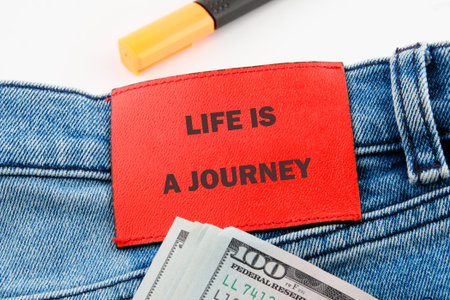 Close up shows a red label on denim jeans with text Life is a Journey. A yellow marker sits atop the jeans and a stack of United States dollars.の写真素材