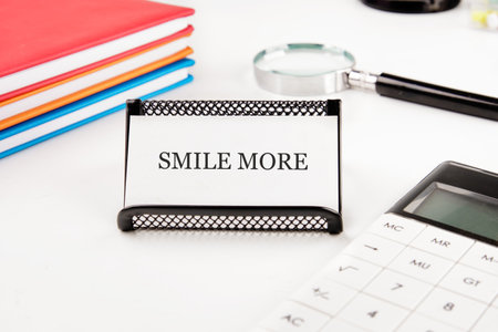 A close up on an office desk. A small sign says SMILE MORE. There are books, a magnifying glass and calculator nearby suggesting a day of work and needing a smile.の写真素材