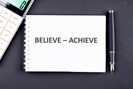 A Notepad with the words BELIEVE - ACHIEVE rests on a dark surface, next to a pen and a calculator, embodying goals, planning and success at Work.の写真素材