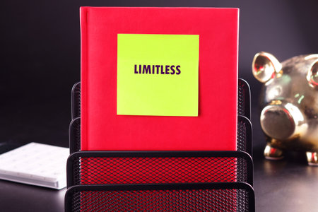 A red notebook with a bright green sticky note reading Limitless sits in a black metal holder. A golden piggy bank is slightly out of focus in the background.の写真素材