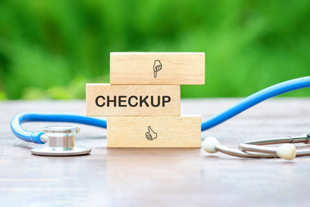 A stethoscope sits beside stacked wooden blocks spelling checkup with icons of a pointing hand and a thumbs up sign, all outdoors against a green, leafy background.の写真素材