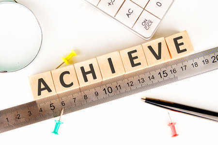 The word ACHIEVE is spelled with wooden blocks across a ruler. A calculator, pen, magnifying glass, and push pins surround the word and measurement tools.の写真素材