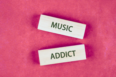On a pink textured surface, two pale wooden blocks display the words Music and Addict in bold green...