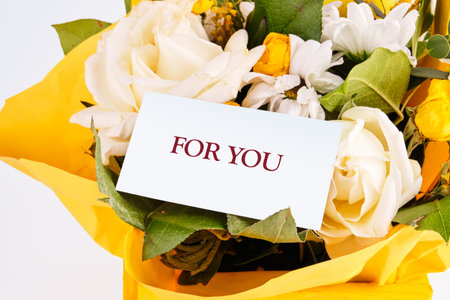 A sunny floral arrangement of white Roses and other flowers presents a card with the words FOR YOU displayed. The gift is wrapped in yellow paper.の写真素材
