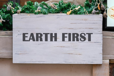 An Earth First sign, in dark letters on distressed white wood, is decorated with garland and pinecones. It hangs outdoors, perhaps near a shop during the daytime.の写真素材