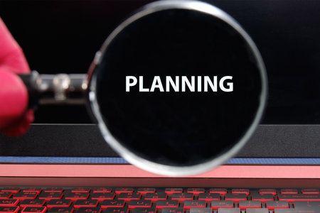 A person uses a magnifying glass to clearly view the word planning on a laptop screen. The glowing keys and dark background create a focused workspace atmosphere.の写真素材