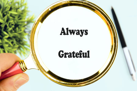 A hand holds a magnifying glass over the always words grateful, symbolizing appreciation and mindfulness in a calm workspace with greenery and a pen nearby.の写真素材