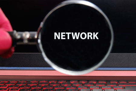 A hand holds a magnifying glass focused on the word NETWORK displayed on a dark laptop screen, symbolizing the exploration of digital connections and technology.の写真素材