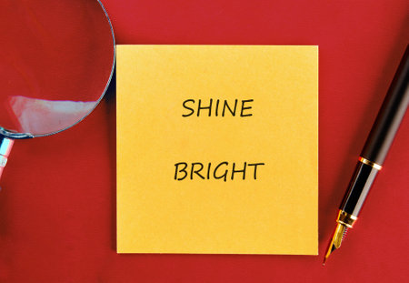 A vibrant yellow sticky note displaying the words shine bright is placed on a deep red surface. Next to it are a sleek pen and a magnifying glass, inviting creativity and inspiration.の写真素材