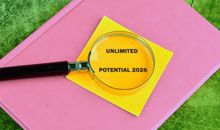 A magnifying glass rests on a bright yellow sticky note that reads unlimited potential 2026, symbolizing focus and clarity, resting on a pink notebook. The settings encourage aspiration.の写真素材