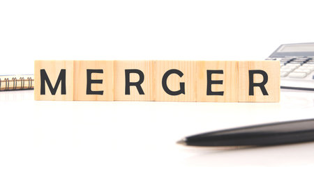 Wooden blocks spell out merger next to a calculator and pen, representing significant business decisions and the potential for growth and collaboration in the corporate landscape.の写真素材