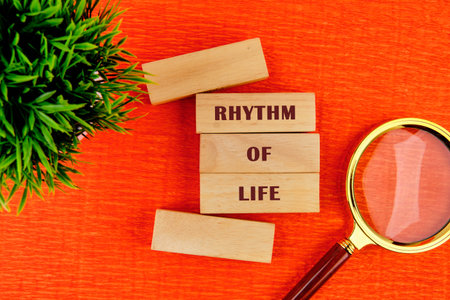 Wooden blocks spell out rhythm of life on a bright orange surface, accompanied by a magnifying glass and a small green plant, capturing the essence of reflection and observation.の写真素材