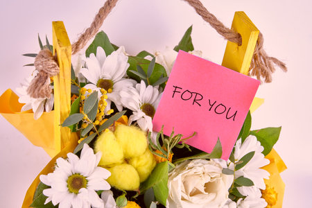 A cheerful bouquet of white daisies, yellow blooms, and greenery is arranged in a vibrant yellow basket. A pink note reading for you adds a personal touch, perfect for gifting.の写真素材