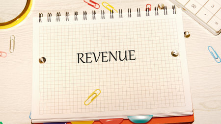 A grid paper notebook displays the word revenue in bold letters. Colorful paperclips and a calculator surround it on a clean, bright workspace, evoking productivity and organization.の写真素材