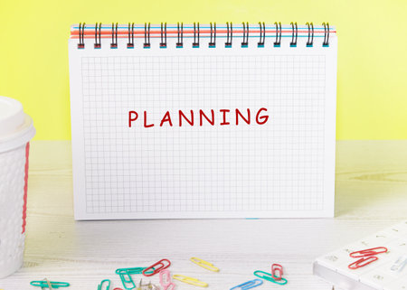 A notebook with the word planning in bold red letters sits on a light wooden surface surrounded by...