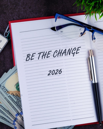 On a desk, the phrase Be the change stands out on a notepad, surrounded by cash, a pen, glasses, and a small plant, symbolizing hope for 2026 and personal growth.の写真素材