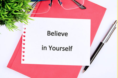 A vibrant workspace is shown with a bright pink notebook, a stylish pen, and a card that reminds us to believe in ourselves. Green plant adds a fresh touch to the setup.の写真素材