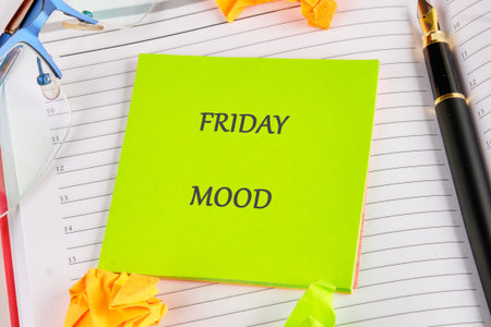 A vibrant green sticky note with the words Friday mood stands out on a tidy desk. Surrounding it are crumpled papers, a pen, and glasses, creating a lively office atmosphere.の写真素材