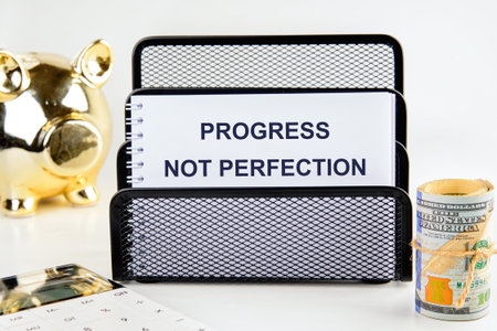 A gold piggy bank, a calculator, and a roll of cash sit near a metal organizer holding a card that reads Progress Not Perfection, against a plain backdrop in a brightly lit setting.の写真素材