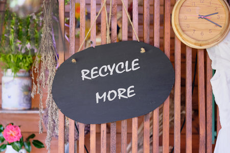 A black wooden sign hangs in a vibrant garden, encouraging people to recycle more. Surrounding greenery and flowers create an inviting and eco-friendly atmosphere. A wooden clock adds charm.の写真素材
