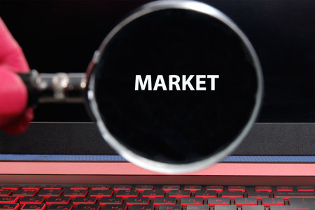 A hand holds a magnifying glass over a laptop screen displaying the word market. This scene captures the essence of searching and analyzing online business opportunities.の写真素材