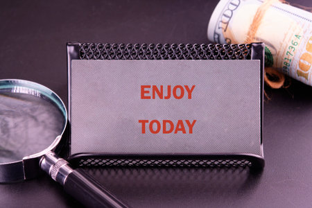 The words ENJOY TODAY are displayed prominently in orange against a gray background. A rolled stack of money sits to the right, and a magnifying glass rests to the left.の写真素材