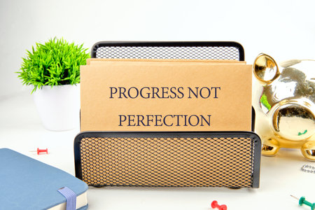 A kraft paper card rests in a black wire holder. Inscribed are the words PROGRESS NOT PERFECTION....